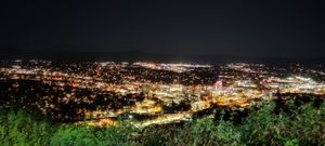 A view of downtown Roanoke from the overlook point at Mill Mountain. The city’s lights are bright and colorful, and the view seemingly goes on forever. The green tops of some trees are in the foreground, with the dark night sky in the background.
