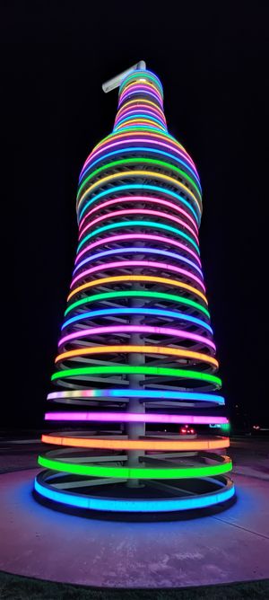 a very tall large soda bottle lit up against the dark sky; there are rainbow-colored rings starting at the bottom and leading all the way to the top, where there is a giant straw