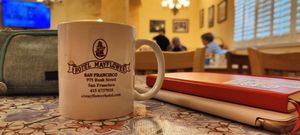 a table in the dining room of the hotel, with my iPad and notebook stacked on the right, and a coffee mug on the left. The coffee mug is branded with Hotel Mayflower and its information: 975 Bush Street, San Francisco, 415 673 7010, sfmayflowerhotel.com