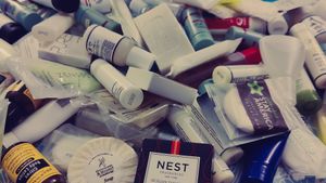 a side view of a collection of toiletries collected from various hotels, spread out on a table, including bar soap, body wash, shampoo, conditioner, hand lotion, body lotion, sewing kits, and hair caps