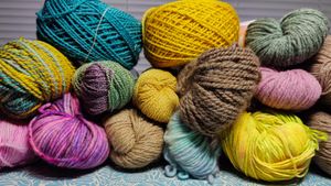 A side view of a pile of yarn skeins, in a variety of colors, weights, and fibers. The pile includes 13 skeins and 2 cakes of yarn. The colors range from vibrant teals to muted tans, and the fibers include wool, alpaca, and more. They range in weight from fingering to aran. Each of the yarns is described in more detail in the blog post.